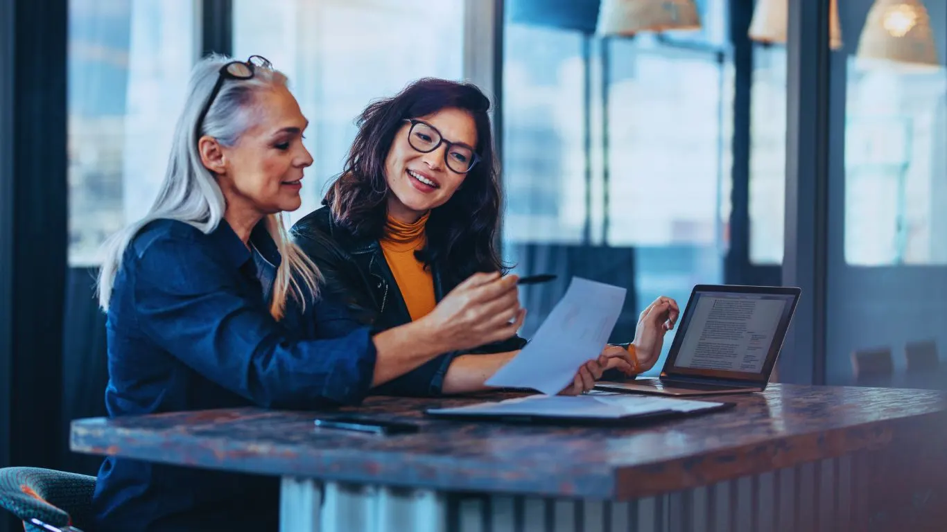 Two professional women collaborating on EU Pay Transparency Directive compliance, reviewing documents and working together on a laptop in a modern office environment